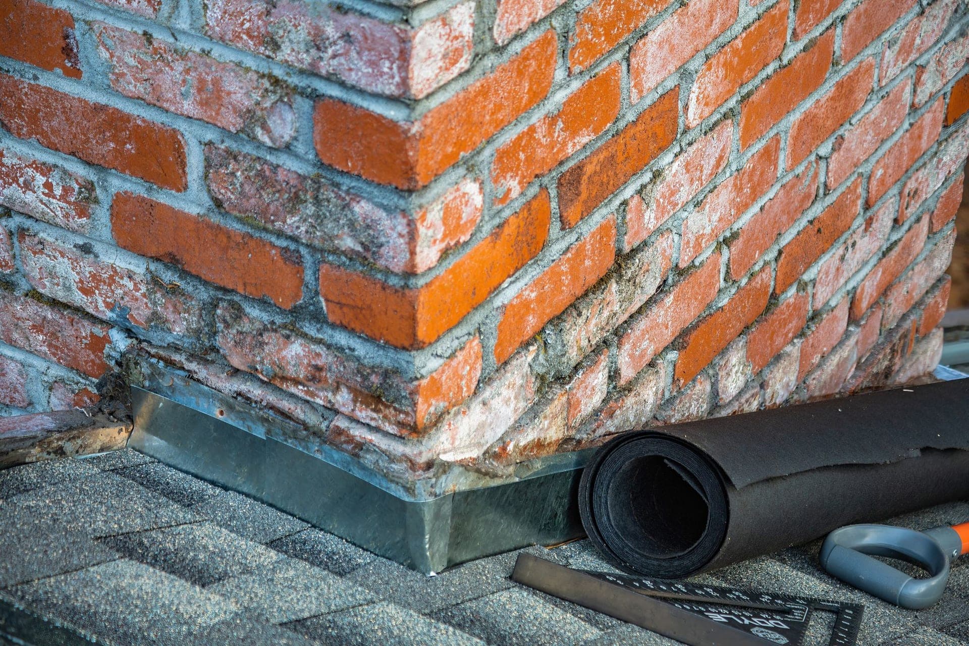 Brick chimney base with new step flashing, a roll of underlayment and a square on the shingles